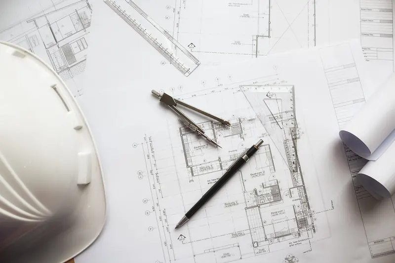 مخططات معمارية على طاولة العمل مع خوذة هندسية وفرجار ومسطرة